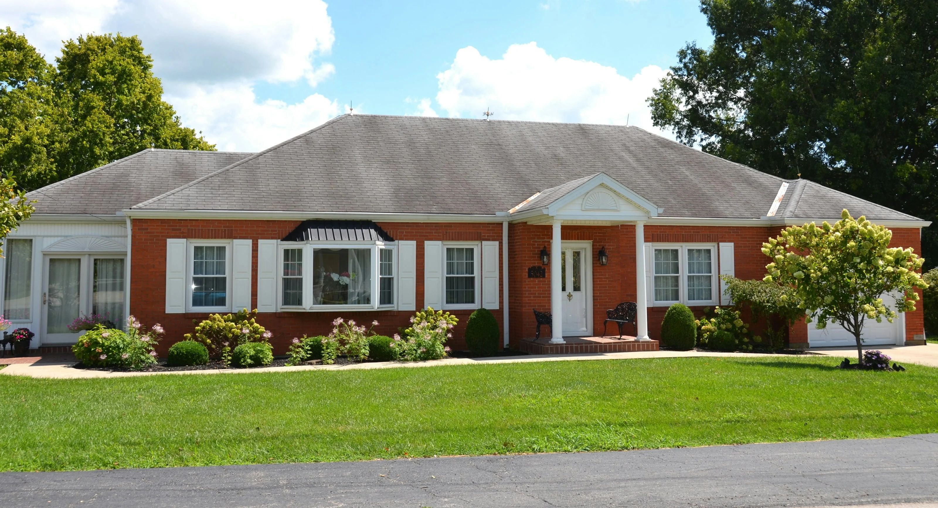 A single-story brick ranch home with mature landscaping and a wide green lawn, seen from the driveway